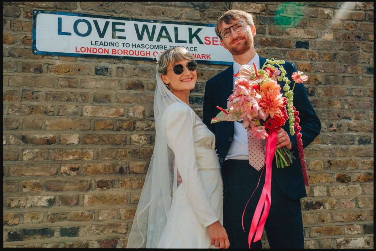 Bride in a veil and sunglasses stands beside her bearded groom holding a bright pink bouquet under the Love Walk street sign