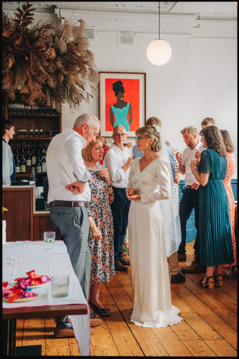 Guests mingle on the wooden floor beneath a dried flower installation and bold portrait, bride chatting in the middle
