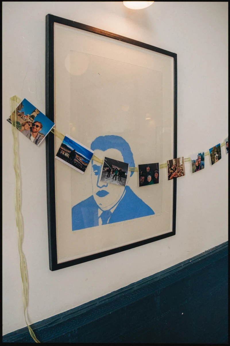 Small printed photos strung on pale green ribbon across a framed blue portrait print at Camberwell Arms