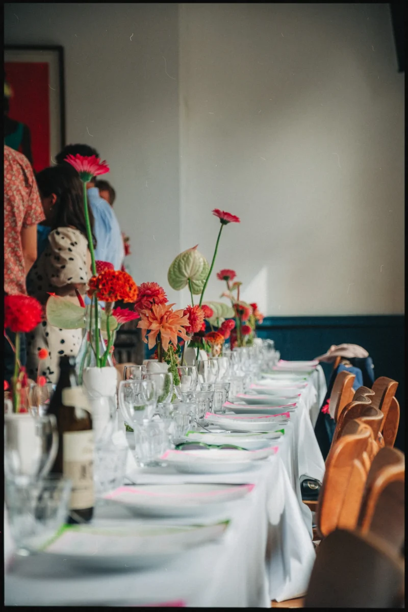 Long white-clothed table laid with plates and glasses, bright zinnias and dahlias in vases, guests standing beyond