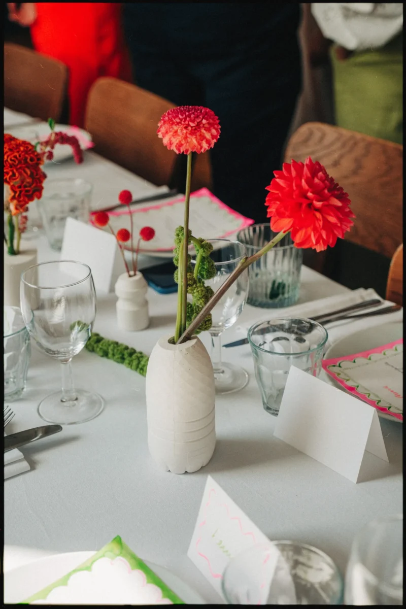 Red and coral dahlias in a white bottle-shaped vase on a laid wedding table with blank place cards and neon pink menus
