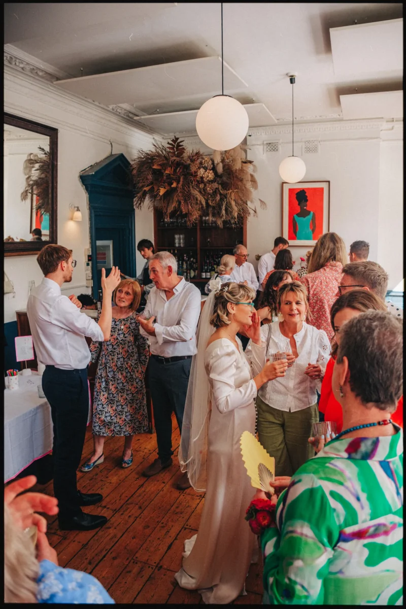 Bride in a veil and long-sleeved white dress chats with guests holding drinks in the upstairs room at Camberwell Arms
