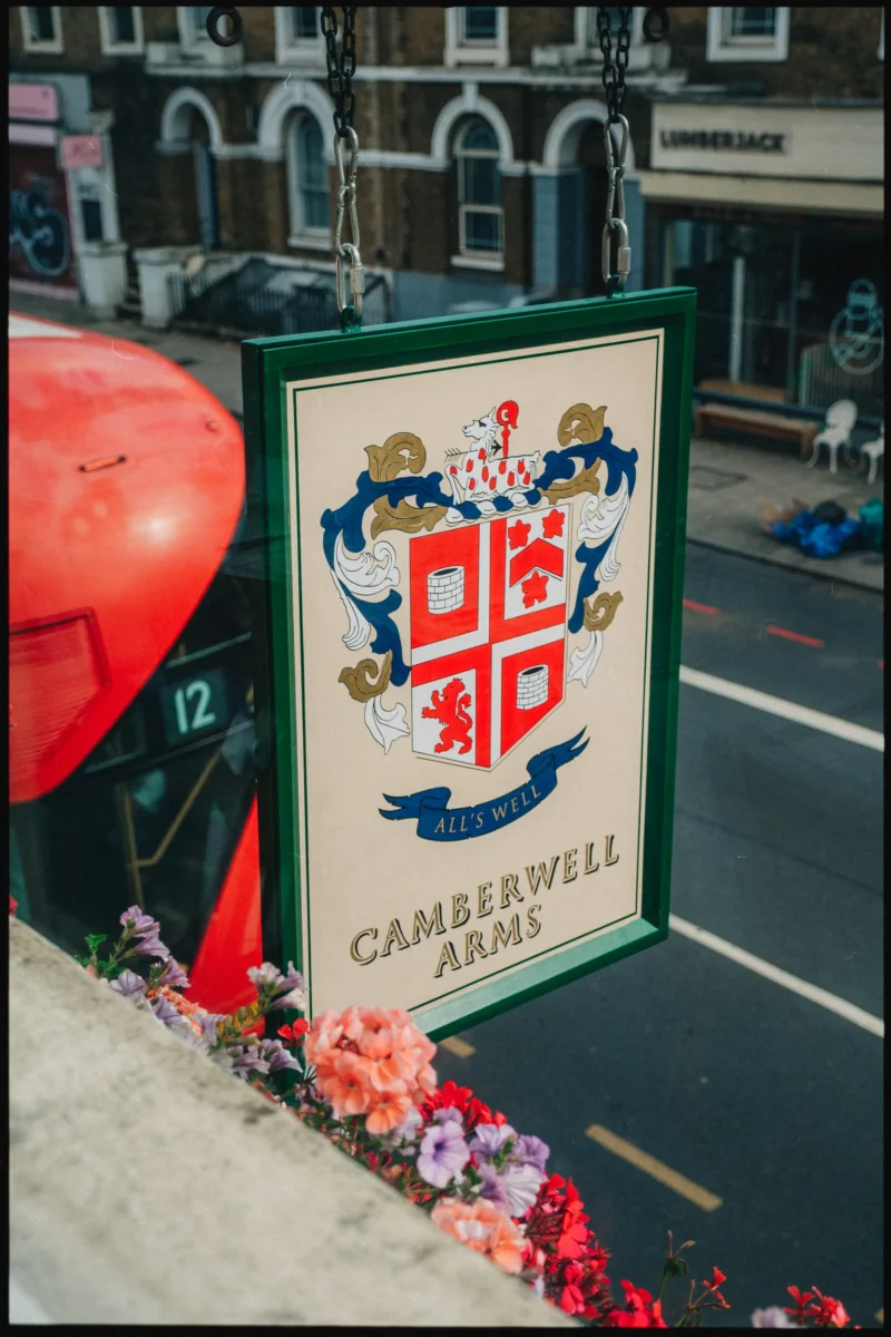 Hanging green pub sign with a crest and the motto All's Well above a London street, a red bus and flowers below