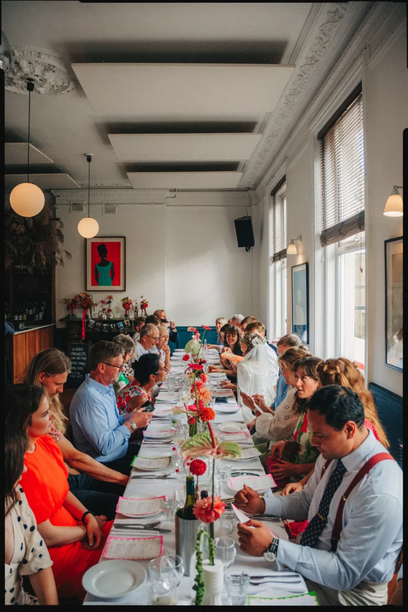 Guests chat and read menus along a long lunch table with bright pink and red flowers in the upstairs room at Camberwell Arms