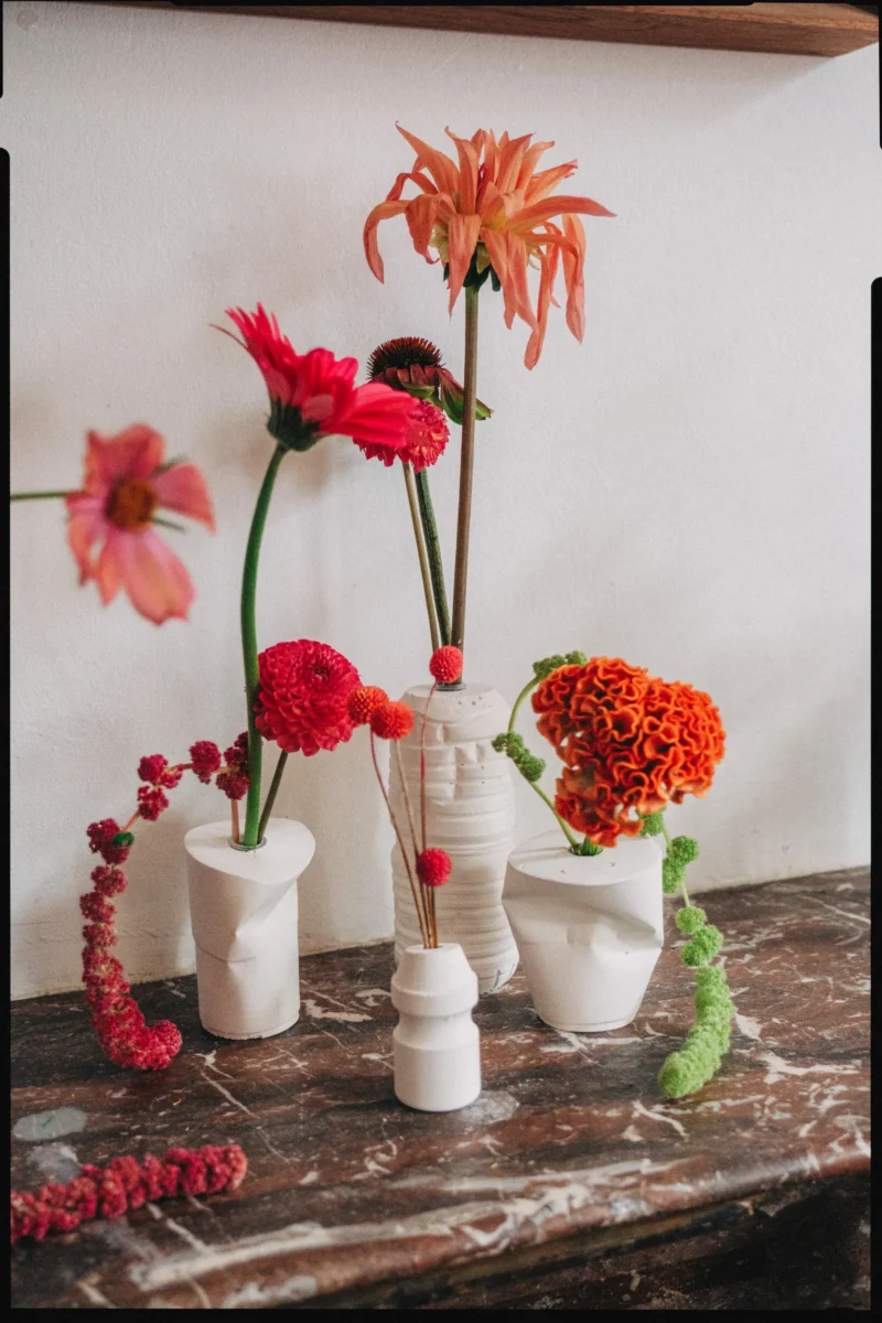 Orange and red dahlias, cockscomb and trailing amaranthus in white sculpted vases on a marble mantelpiece