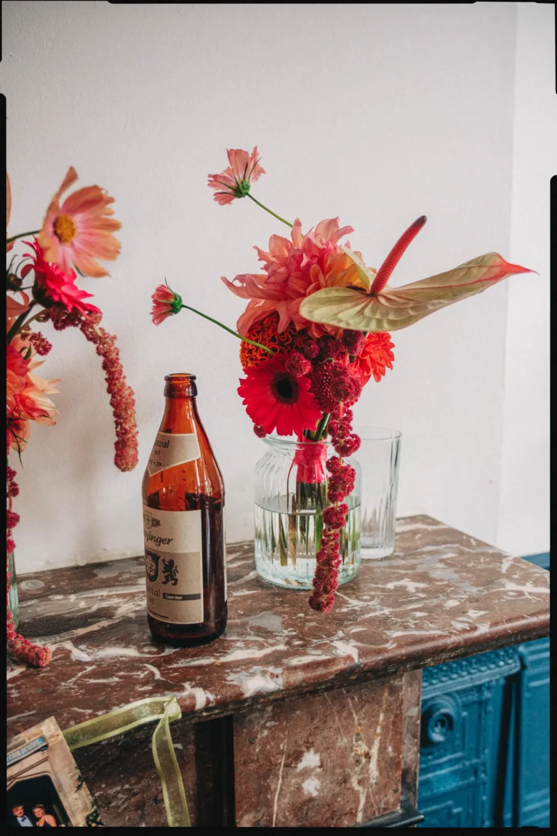 Bold red and coral dahlias, anthurium and trailing amaranthus in a glass jar beside a beer bottle on a marble mantelpiece