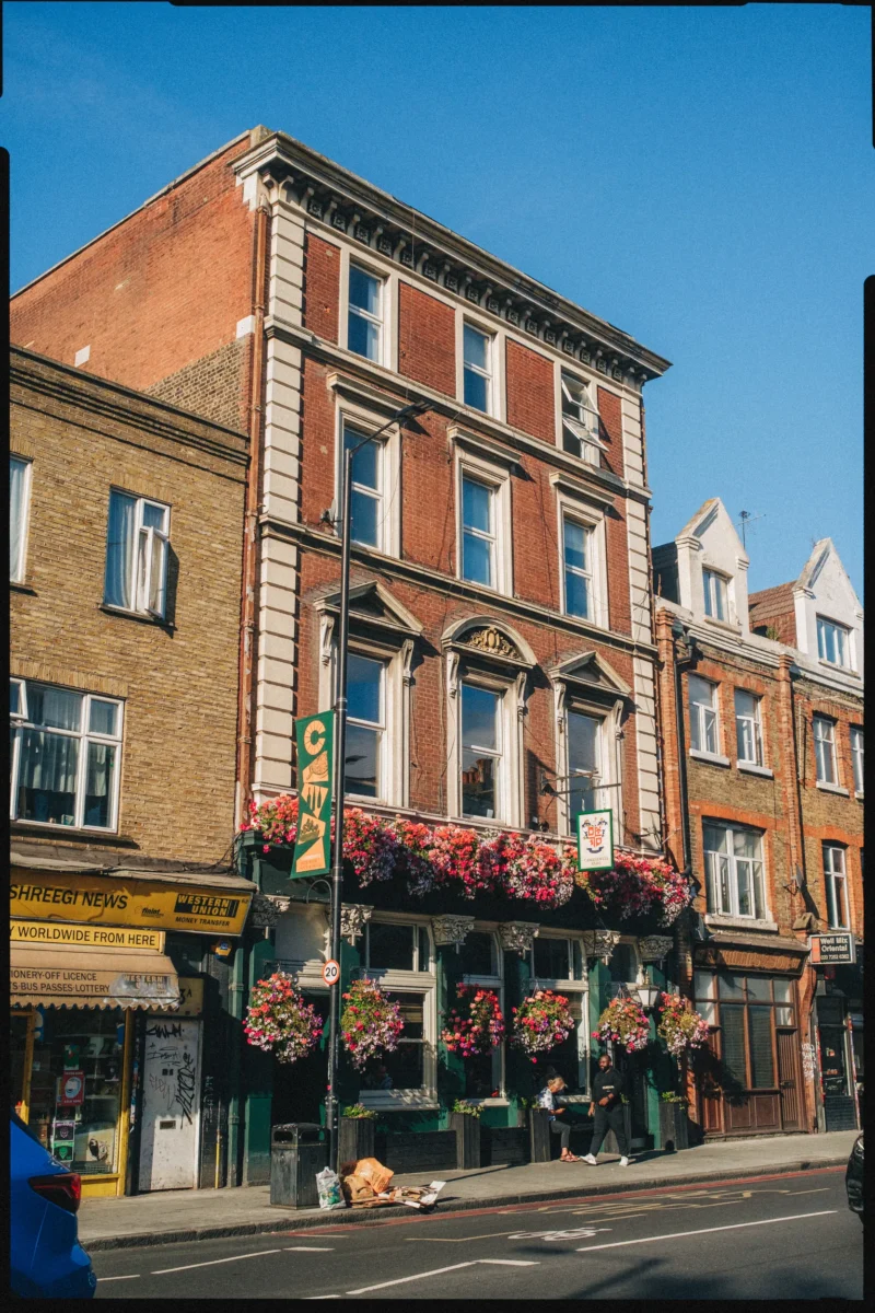 Red-brick Camberwell Arms pub front with pink hanging baskets under a blue summer sky, two people on the pavement