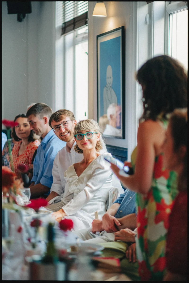 Bride in teal glasses beams along the table as a guest stands to speak during lunch at Camberwell Arms