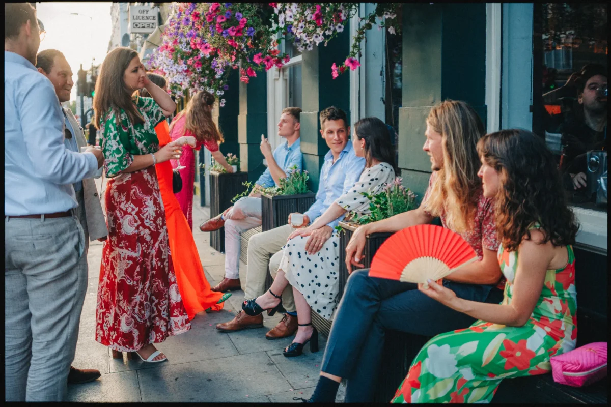 Guests in summer dresses chat on the pavement outside Camberwell Arms, one woman holding a red paper fan