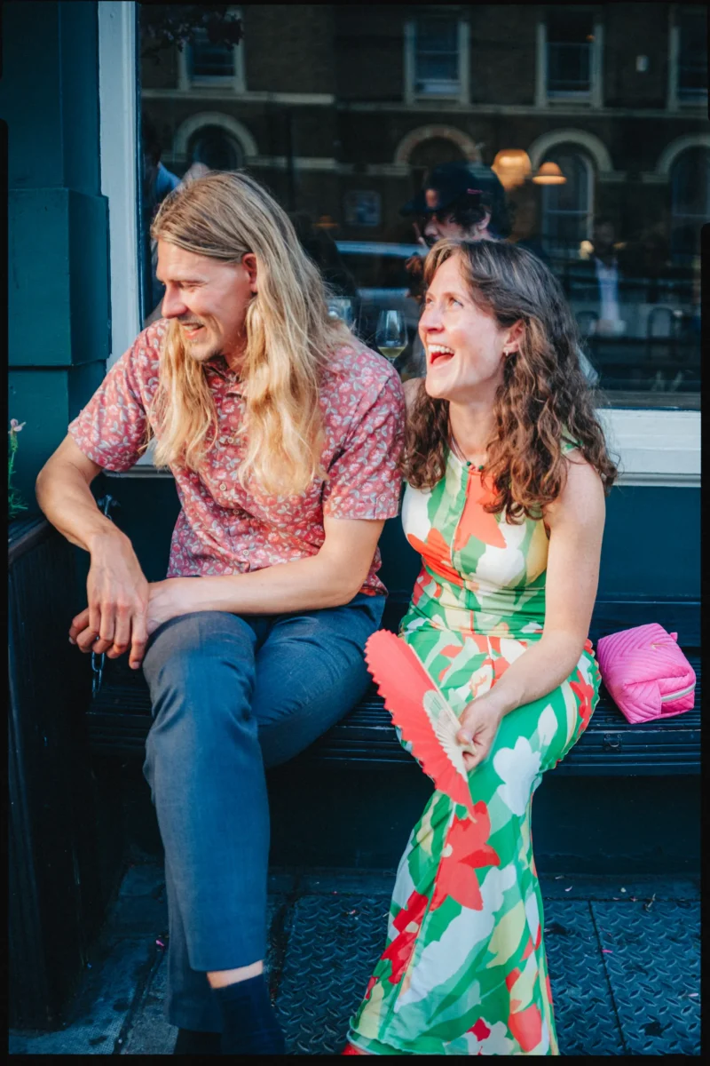 Laughing woman with a red fan and a long-haired man in a pink shirt share a bench outside Camberwell Arms