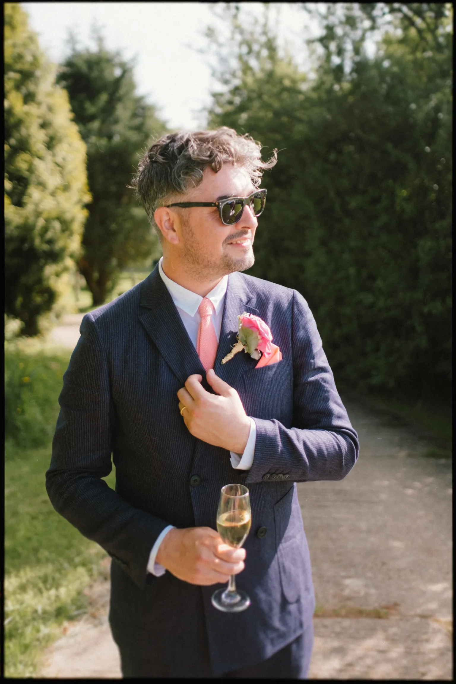 A man in a navy suit with a pink tie and boutonnière stands outdoors at a Yoghurt Rooms wedding festival, holding a glass of champagne and wearing sunglasses, looking off to the side with a smile.
