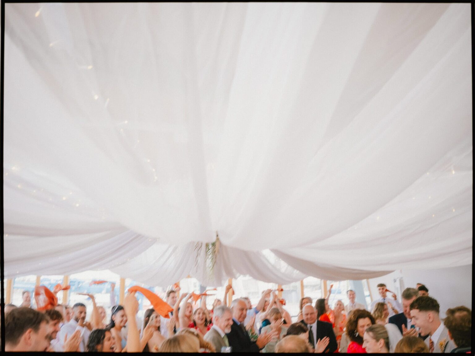 Napkins in the air and cheers all around — the bride and groom make their entrance to the wedding breakfast at Greenwich Yacht Club in London, captured beneath flowing white drapes. Guests waving napkins as the bride and groom arrive for the wedding breakfast at Greenwich Yacht Club in London, captured beneath flowing white ceiling drapes.