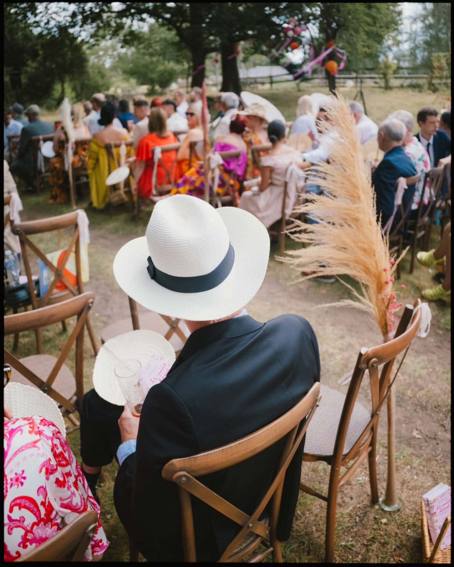 A stylish guest in a sun hat enjoys the outdoor ceremony at Guy’s Cliffe House in Warwickshire, surrounded by guests and rustic dried flower details. Wedding guest in a sun hat sitting at the outdoor ceremony with dried flower decorations at Guy’s Cliffe House in Warwickshire.