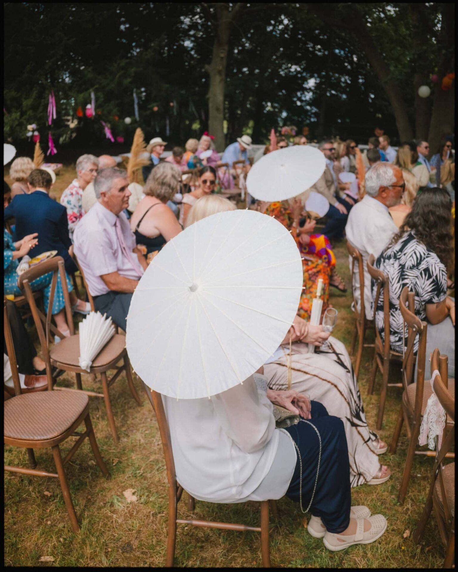 Guests shade themselves with parasols during a sunny outdoor ceremony at Guy’s Cliffe House in Warwick — a relaxed summer wedding moment. Guests sitting with white parasols during a sunny outdoor wedding ceremony at Guy’s Cliffe House in Warwick.