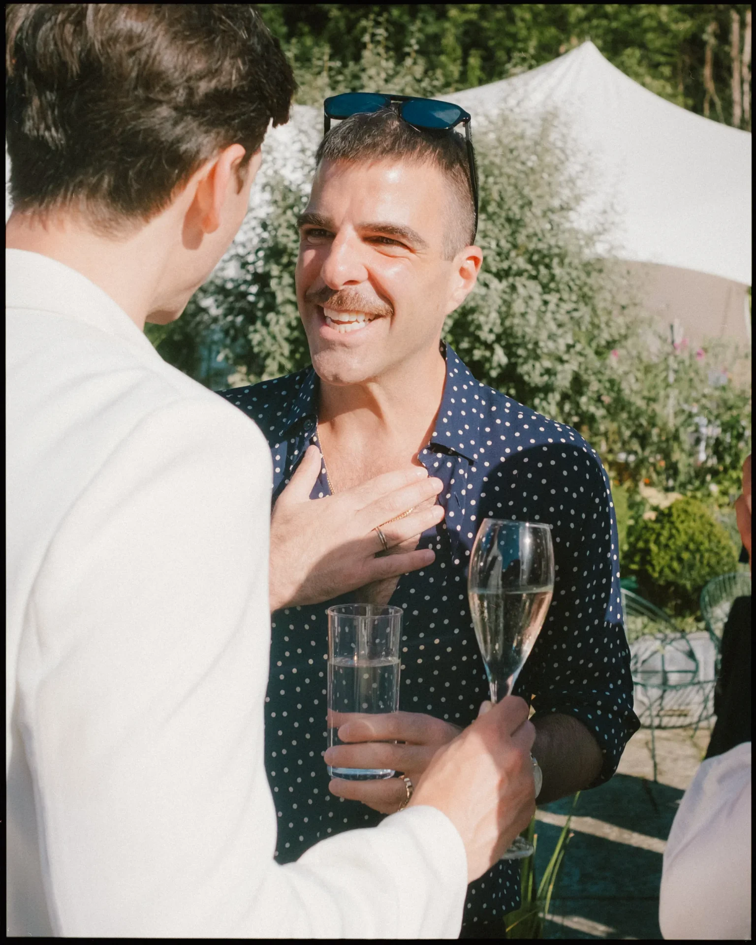 A man in a navy blue polka-dot shirt smiles while holding a glass of champagne and talking to another person at an outdoor event. Sunlight highlights their faces and greenery is visible in the background.