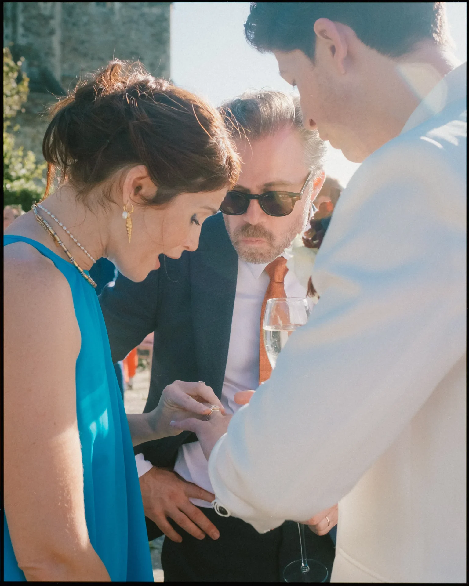 Three people are gathered closely together outdoors. A woman in a blue dress is showing something to two men, one in a dark suit with sunglasses and another in a white jacket, while sunlight streams in from the side.