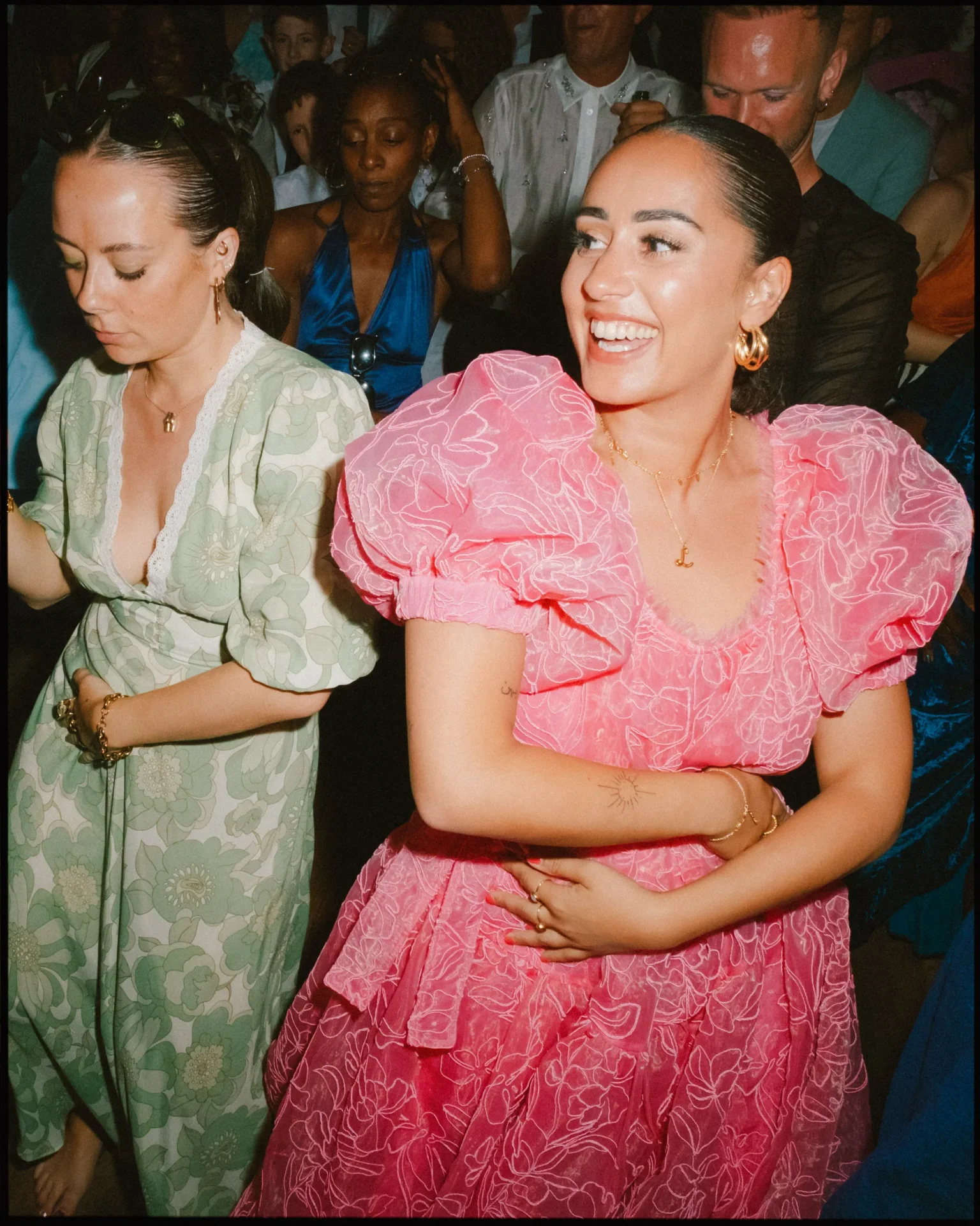 Two women in colorful, elegant dresses dance joyfully at a crowded indoor event. One wears a green floral dress, the other a bright pink dress with puffy sleeves, both smiling and surrounded by other people.