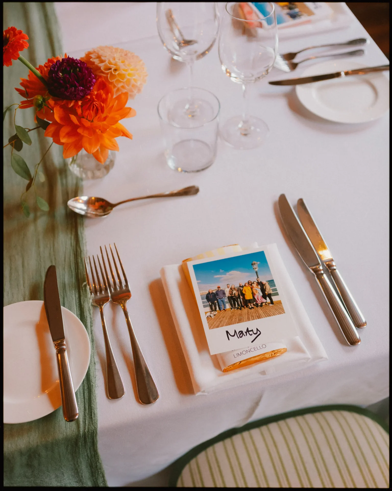 A set dining table with orange and white flowers in a vase, cutlery, empty glasses, and a place card labeled Mary with a group photo, placed on a white napkin and dish atop a white tablecloth.