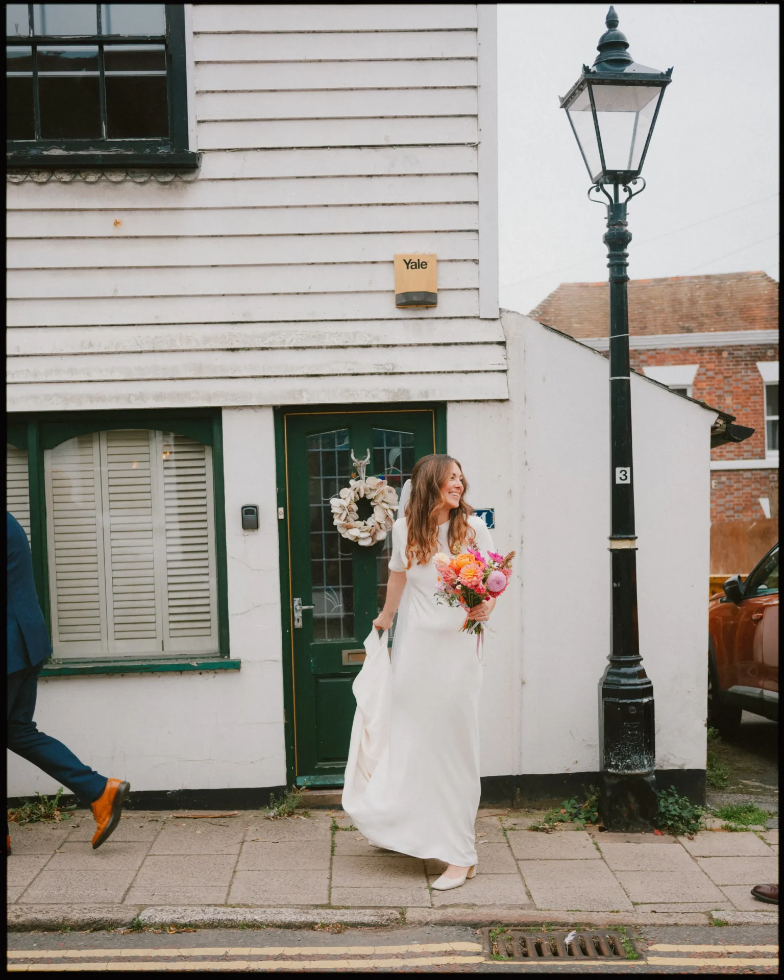 A bride in a white dress holding a colorful bouquet stands smiling on a sidewalk outside a white house; a man in a suit is walking out of the frame on the left. A streetlamp and a parked car are visible nearby.