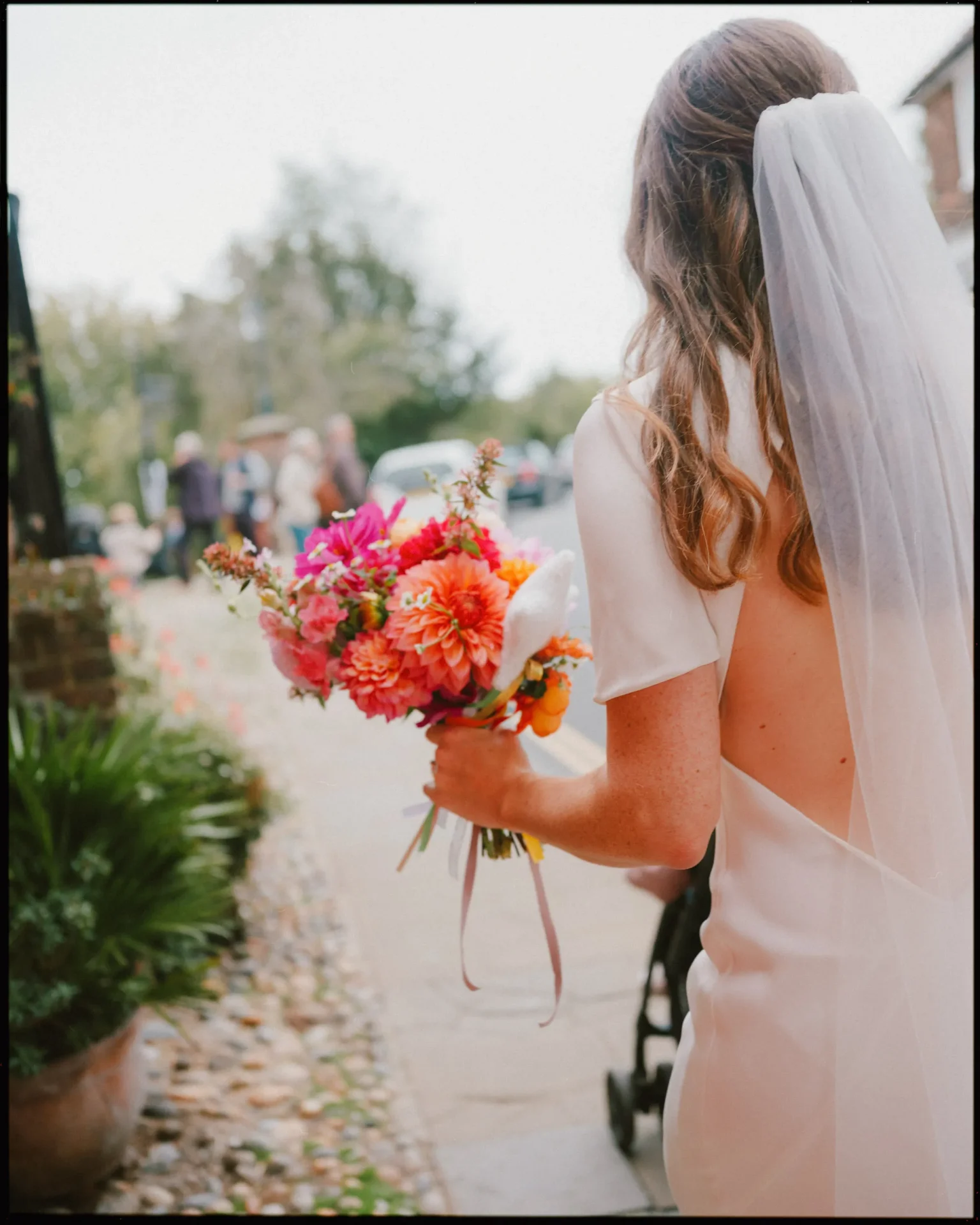 A bride in a white dress and veil holds a colorful bouquet of flowers while walking outdoors on a stone path, with greenery and blurred people in the background.