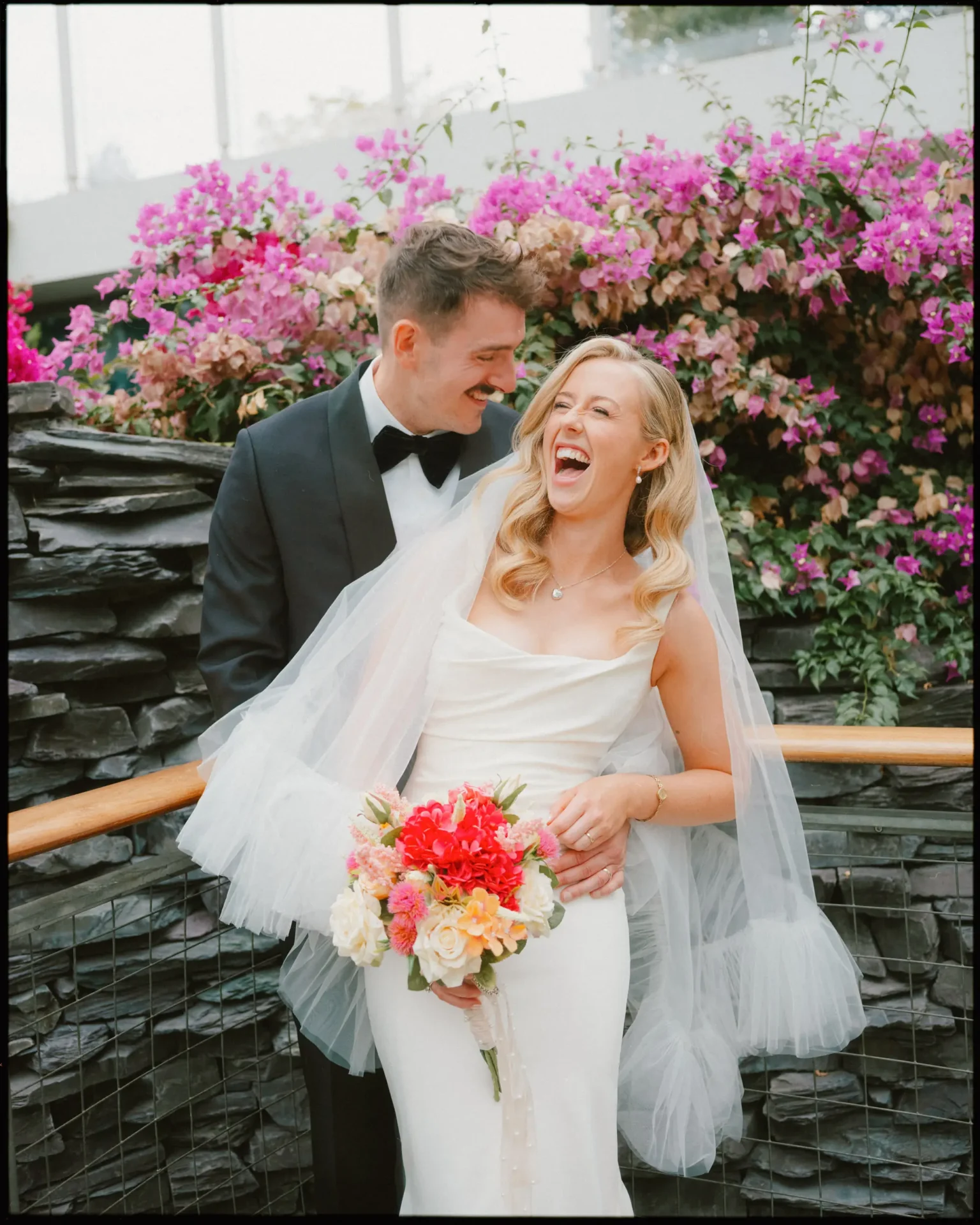 A bride and groom smile and laugh together in front of a stone wall and vibrant pink flowers. The bride holds a colorful bouquet and wears a white dress and veil; the groom wears a black tuxedo.
