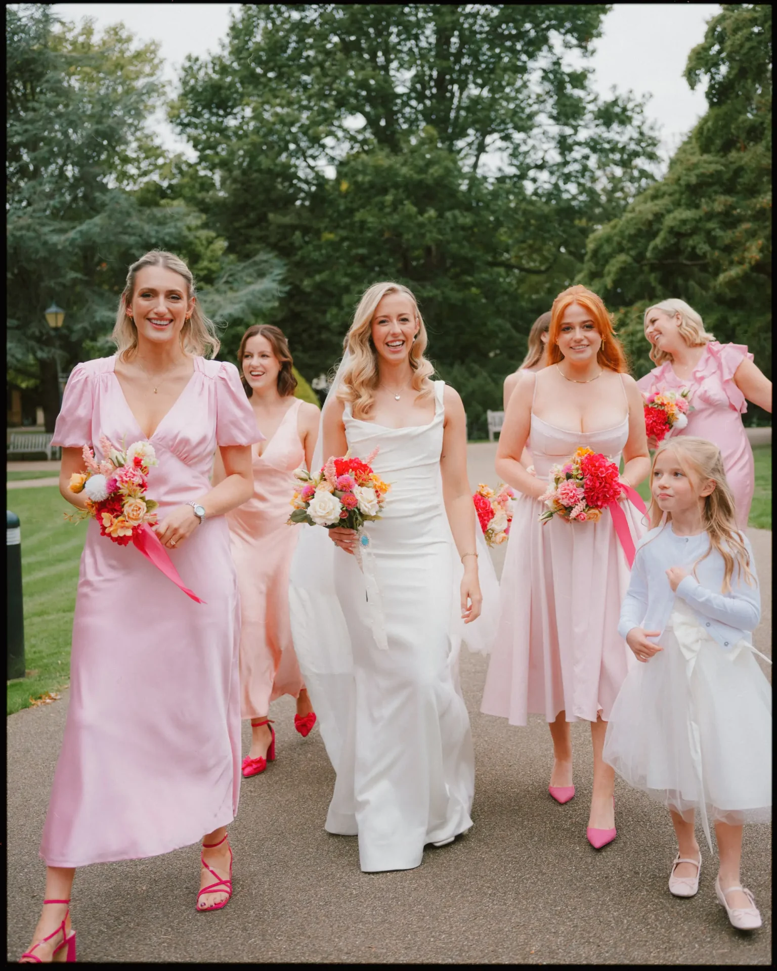 A bride in a white dress walks outdoors with bridesmaids in pink dresses and a young girl in a white dress. They are smiling, holding bouquets, and surrounded by greenery.